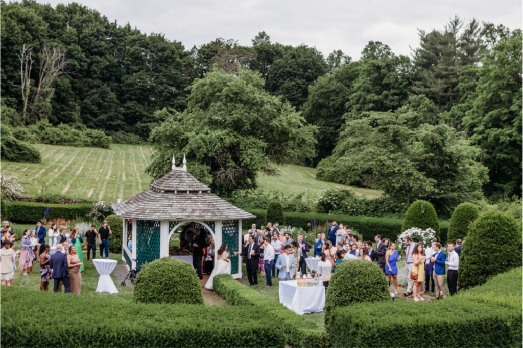 A wide shot of wedding guests gathered around a gazebo in a garden, celebrating after the ceremony, with lush greenery surrounding them.