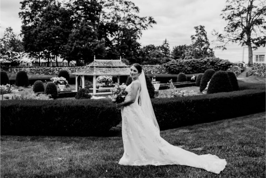 A black and white photo of a bride in a long veil and lace dress holding a bouquet, standing in a manicured garden with a gazebo in the background.