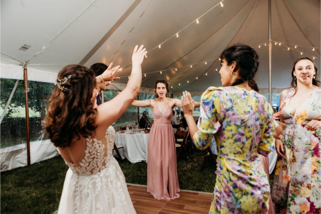 A group of wedding guests dancing under a tent, with string lights overhead. One guest in a pink dress stands in the center with her arms raised, while other guests in floral dresses and a bride in a lace gown join in.