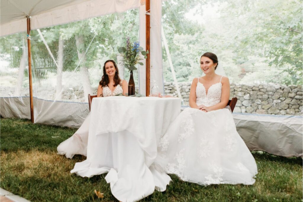 Two brides in white lace wedding dresses seated at a small, round table under a tent. They are smiling and surrounded by greenery visible through the clear tent panels.