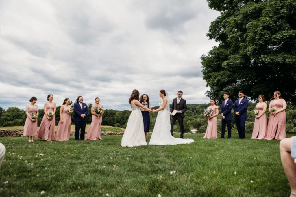 A wedding ceremony taking place outdoors, with two brides holding hands, surrounded by bridesmaids in blush dresses and groomsmen in navy suits.