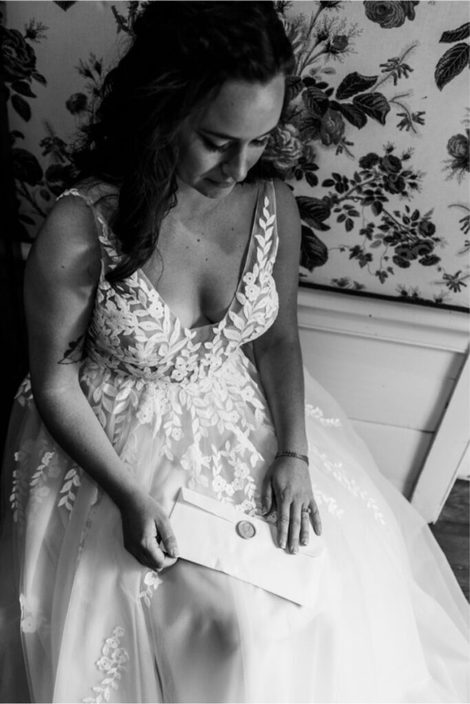 A black-and-white close-up of a bride seated in her wedding gown, holding an envelope with a wax seal, appearing emotional.