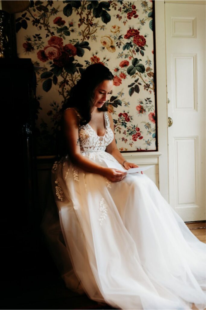 A bride sitting in a floral-wallpapered room, reading a letter with a calm expression, her wedding dress flowing around her.