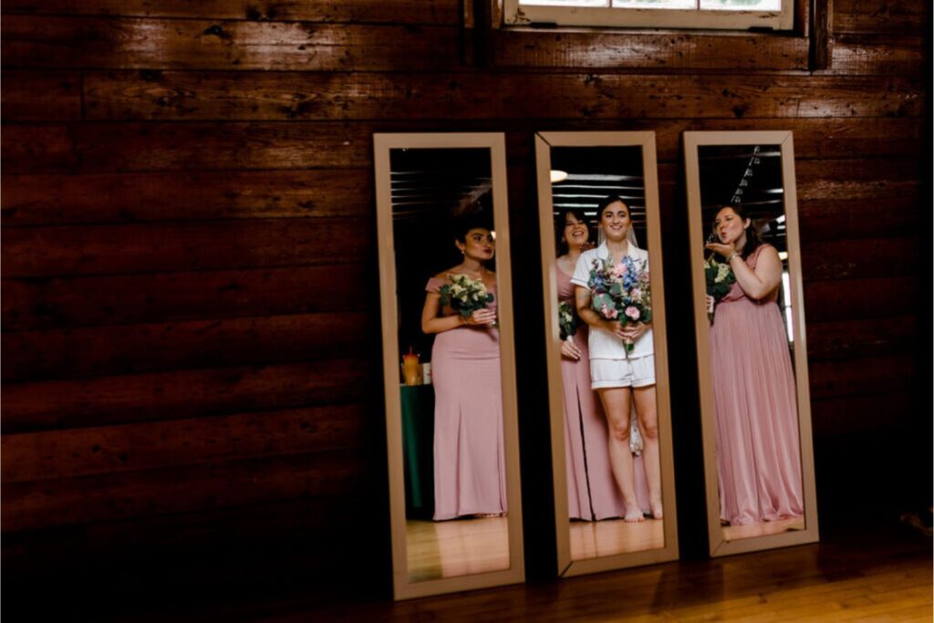 A reflection photo showing the bride and her two bridesmaids posing in front of full-length mirrors inside a wooden cabin.