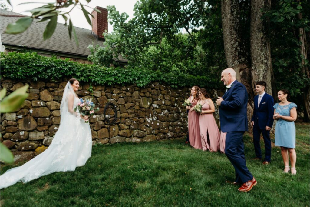 A bride, holding a bouquet, walking towards her father and bridal party, who are waiting with smiles near a stone wall and lush greenery.