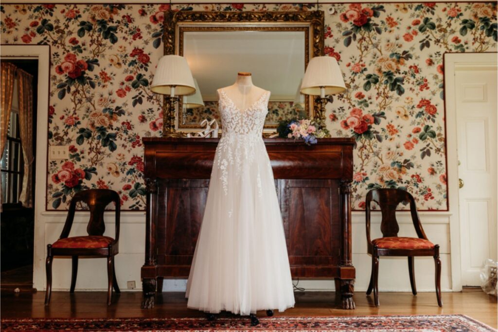 A wedding dress displayed on a mannequin in a vintage room with floral wallpaper and antique furniture.