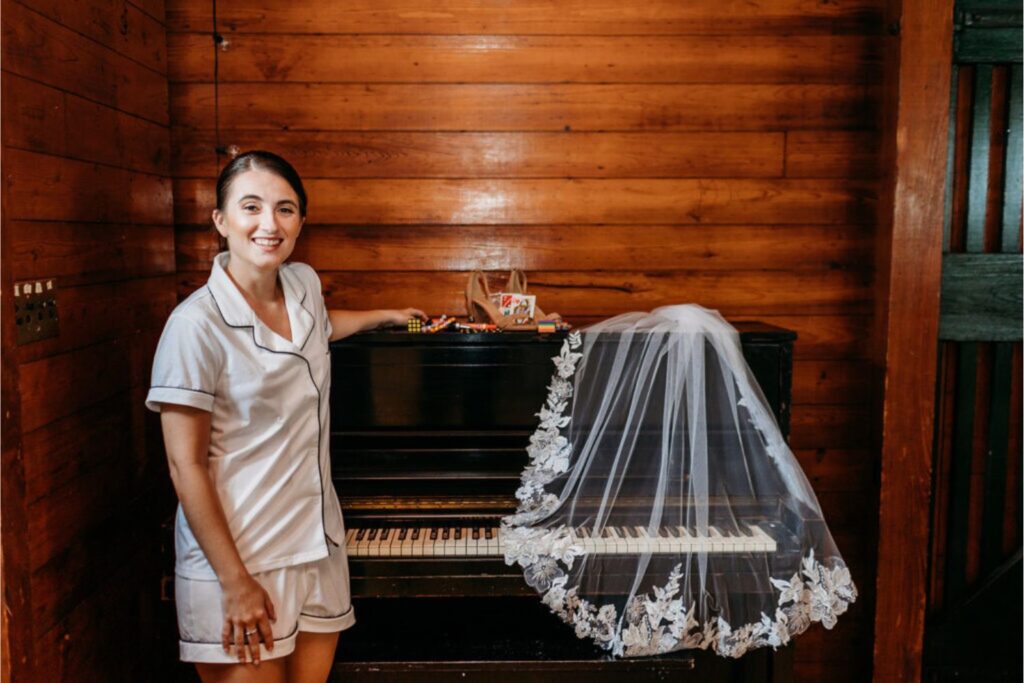 A bride smiling in casual pajamas, standing next to a piano, with her wedding veil draped across the piano keys.