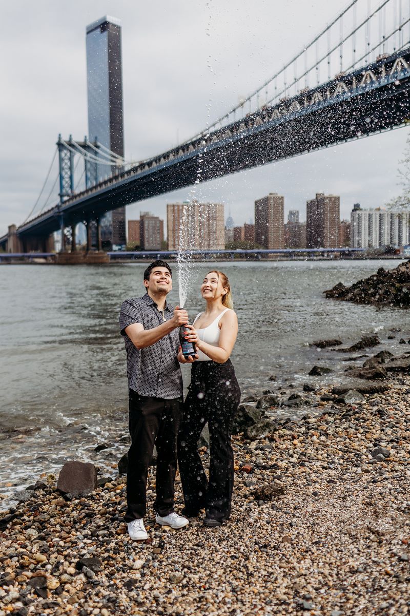 man and woman popping a bottle of champagne at the waterfront under the brooklyn bridge in new york 