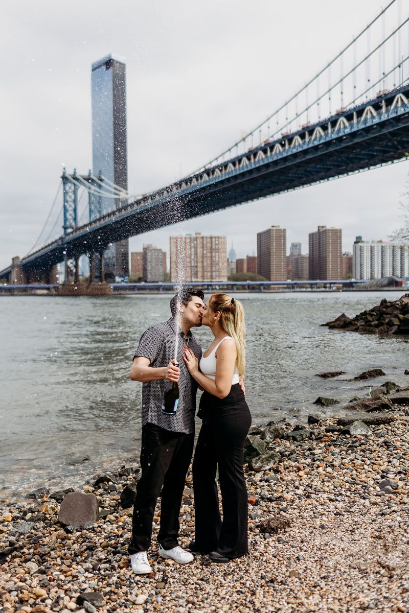 man and woman giving each other a kiss while popping a bottle of champagne underneath the brooklyn bridge 