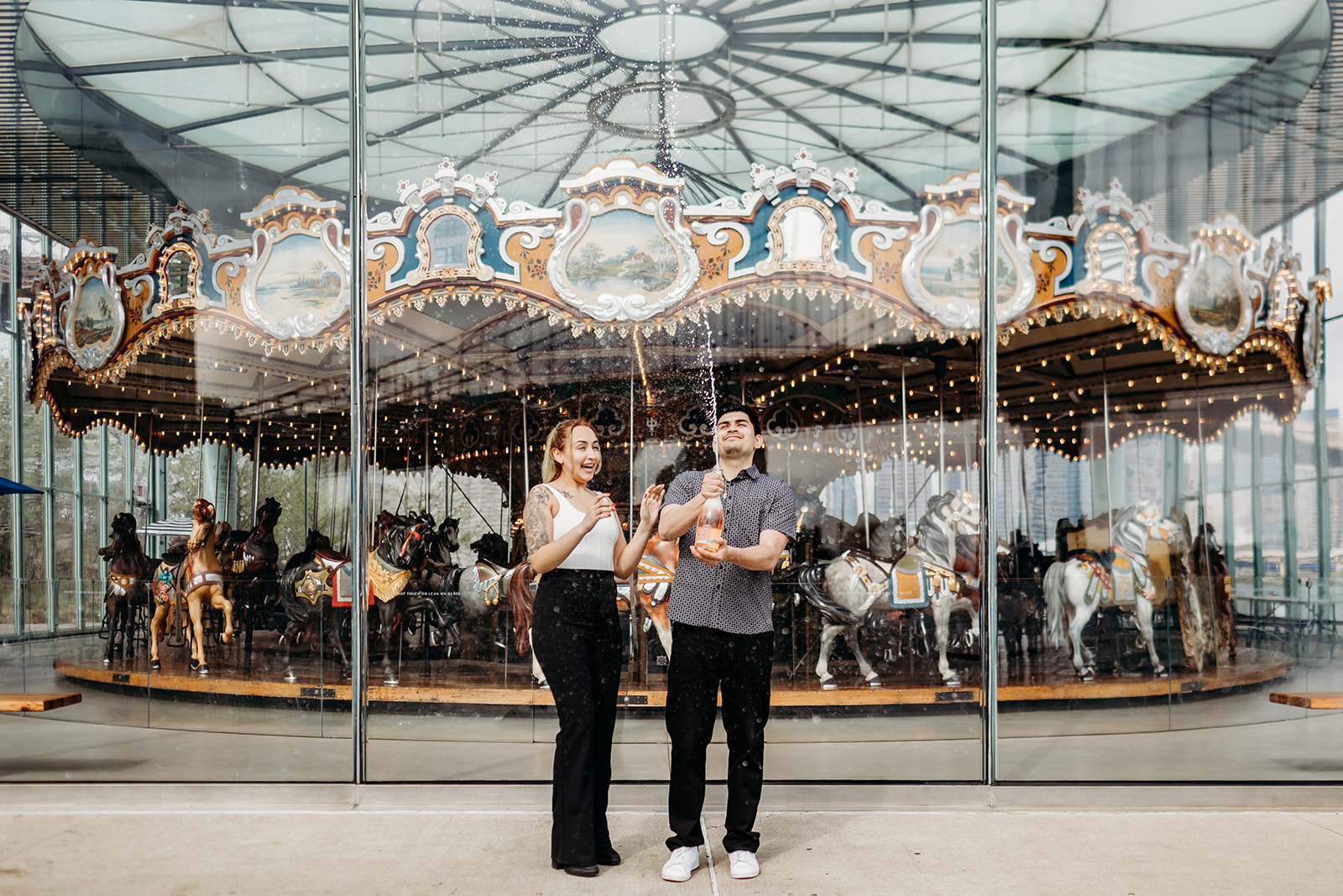 man and woman popping a bottle of champagne in front of a carousel 