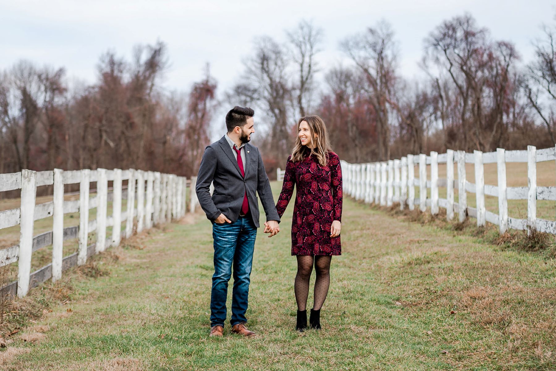 A woman in a red and black dress, tights, and black heels is holding her partner's hand and looking at him and her partner is wearing a gray dress jacket, a red tie, blue jeans, and brown shoes and is looking toward her and both are smiling and around them is a white picket fence and wide open field 