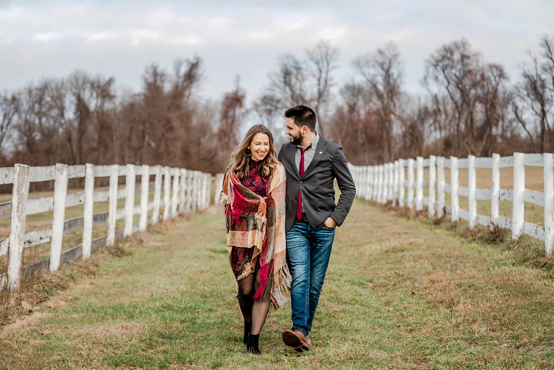 A couple is walking in a field with a white picket fence the woman has a checkered blanket that is red, tan, and green warpped around her and her partner is holding her backside and is wearing a gray suit jacket, blue jeans, brown shoes, and a red tie and is looking towards her