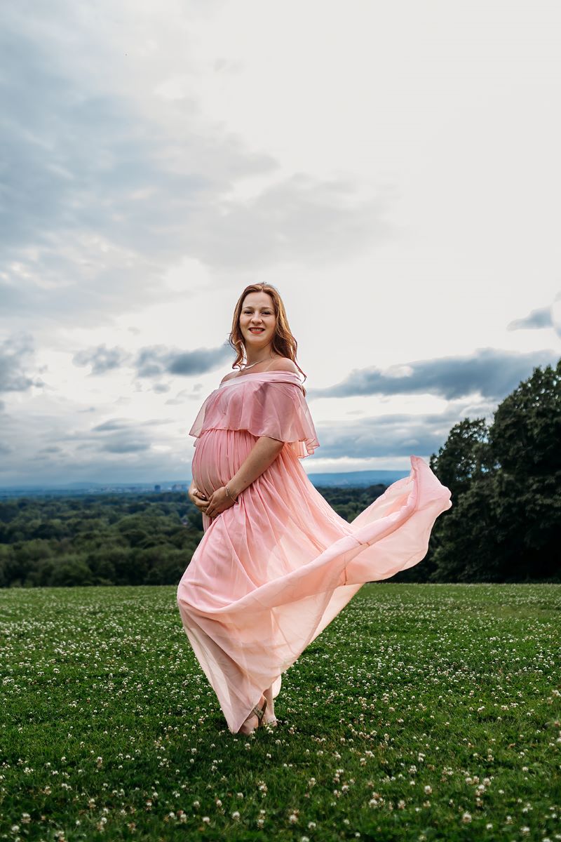 A woman with red hair is wearing a pink dress and she is pregnant and holding her stomach and she is standing in an open field 