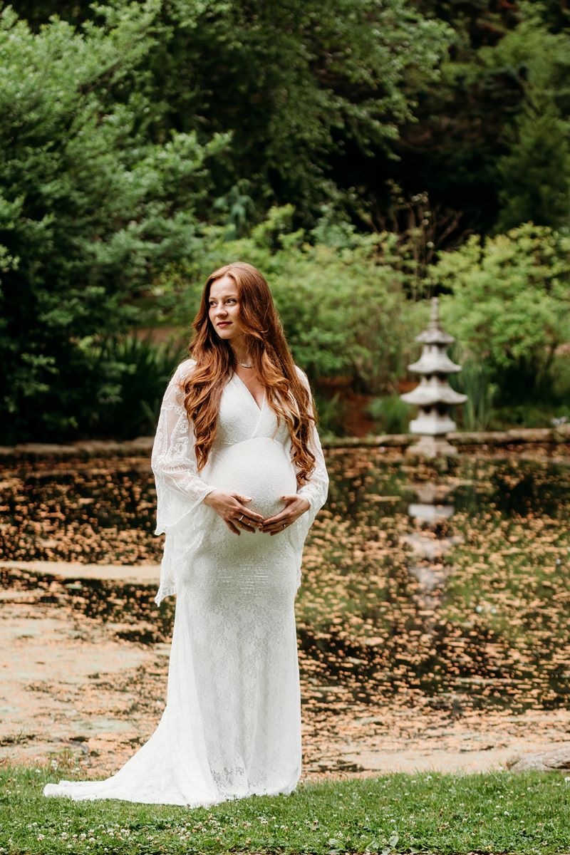 A woman who is pregnant is wearing a white dress and holding her stomach she has red hair and she is looking off to the side 