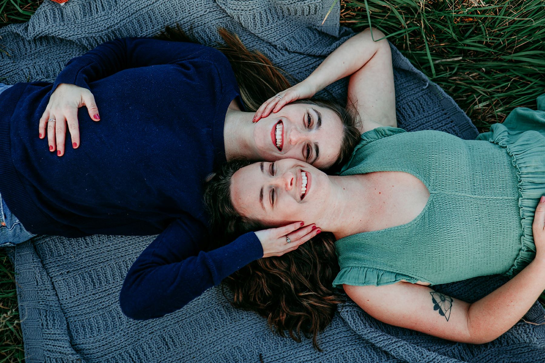 Two women laying on a blue blanket on the gress one woman has on a blue long sleeve shirt and jeans and the other woman has on a mint green short sleeve tops both are using one arm to touch the other's face with their other hand resting on their stomachs and both are smiling 