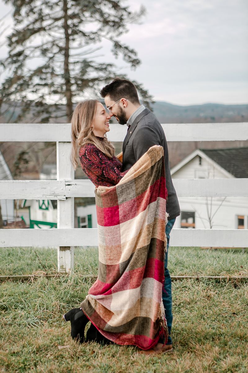 A woman with a red patterned dress is holding a blanket around her arms that is checkered and is red, tan, and green and she is looking at her partner who is wearing a gray suit jacket and jeans and glasses and who is leaning towards her behind them is a white fence 