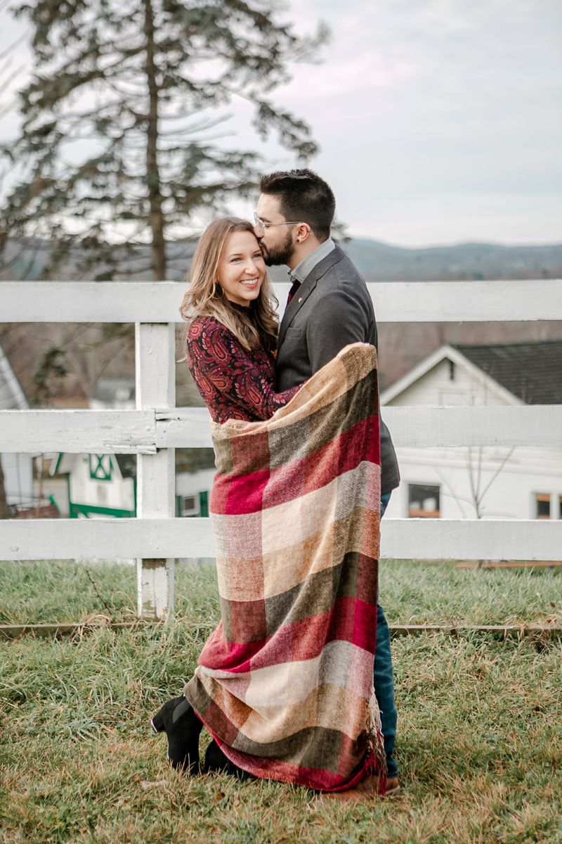 A woman with a red patterned dress is holding a blanket around her arms that is checkered and is red, tan, and green and her partner who is wewaring a gray suit jacket and jeans and glasses is leaning towards her and kissing the side of her forehead behind them is a white picket fence 