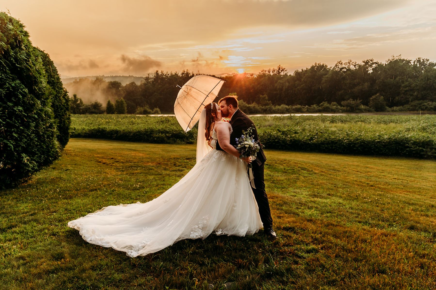 A woman in a white wedding dress and veil is kissing her husband who is wearing a black suit and holding an umbrella over their heads the woman is also holding a bouquet of flowers in her hand that is made up of white and blue flowers they are standing in a field and the sun is setting behind them 