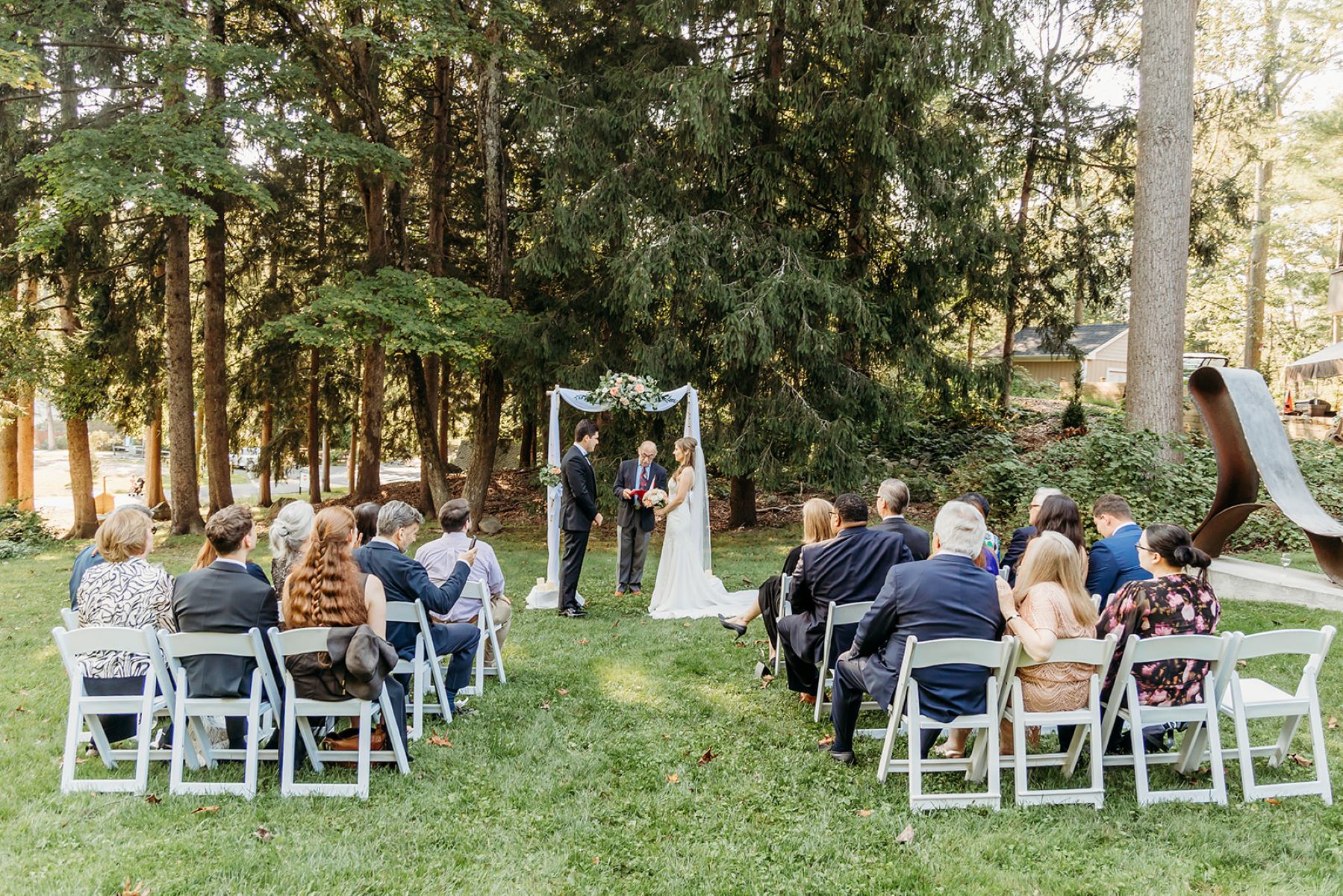 A man and a woman at their micro wedding with their guests who are sitting in white chairs and the woman is wearing a white wedding dress and holding a bouquet of flowers and the man is wearing a black suit and they are both looking at each other bheind them is a white arch and their wedding officiant and they are surrounded by trees, grass, and plants 