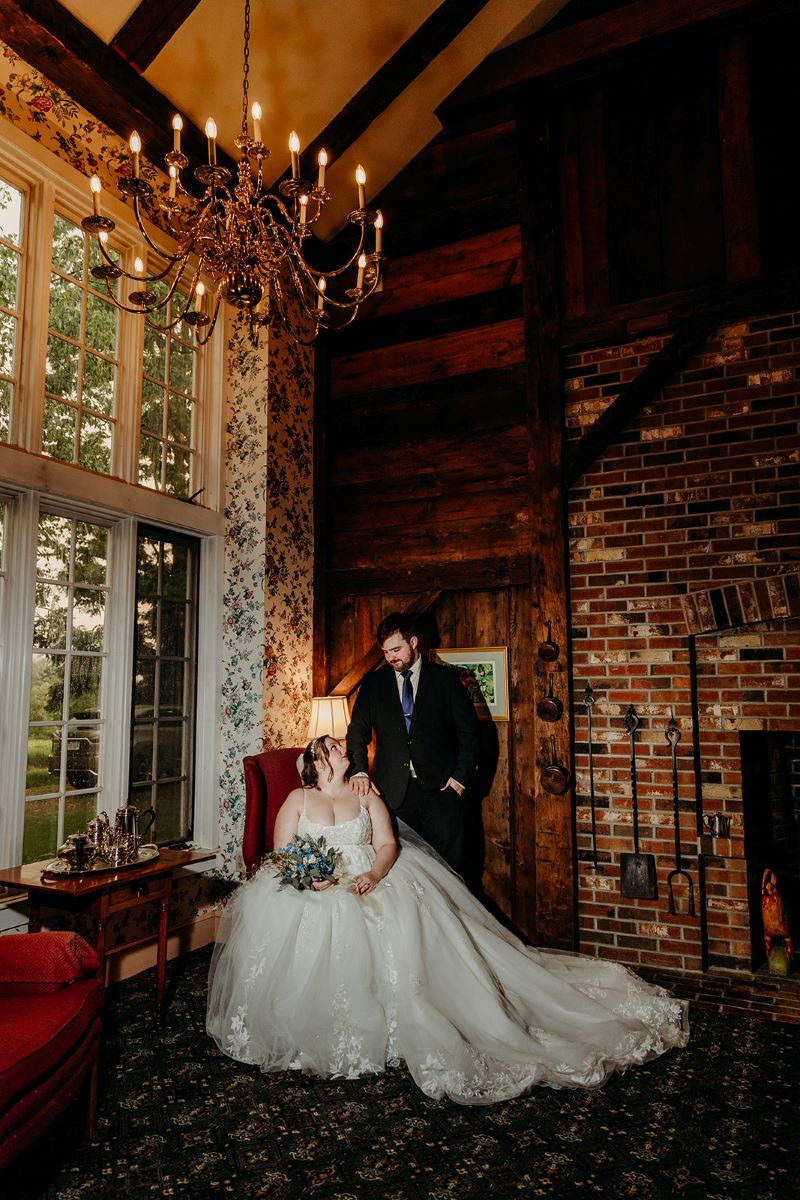 A woman in a white wedding dress is sitting in a red chair and she is holding a bouquet of flowers in her hand made up of green, blue, and white flowers she is looking up at her husband who is touching her shoulder and looking down at her and he is wearing a black suit behind them is a brick wall and a big bay window and a wall with floral wallpaper and a fireplace 