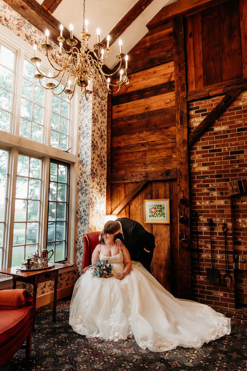 A woman in a white wedding dress is sitting in a red chair and holding a bouquet of flowers in her hands that is resting on her lap and she is kissing her husband who is wearing a black suit and behind them is a large bay window, a table with a tea set, and an exposed brick wall 