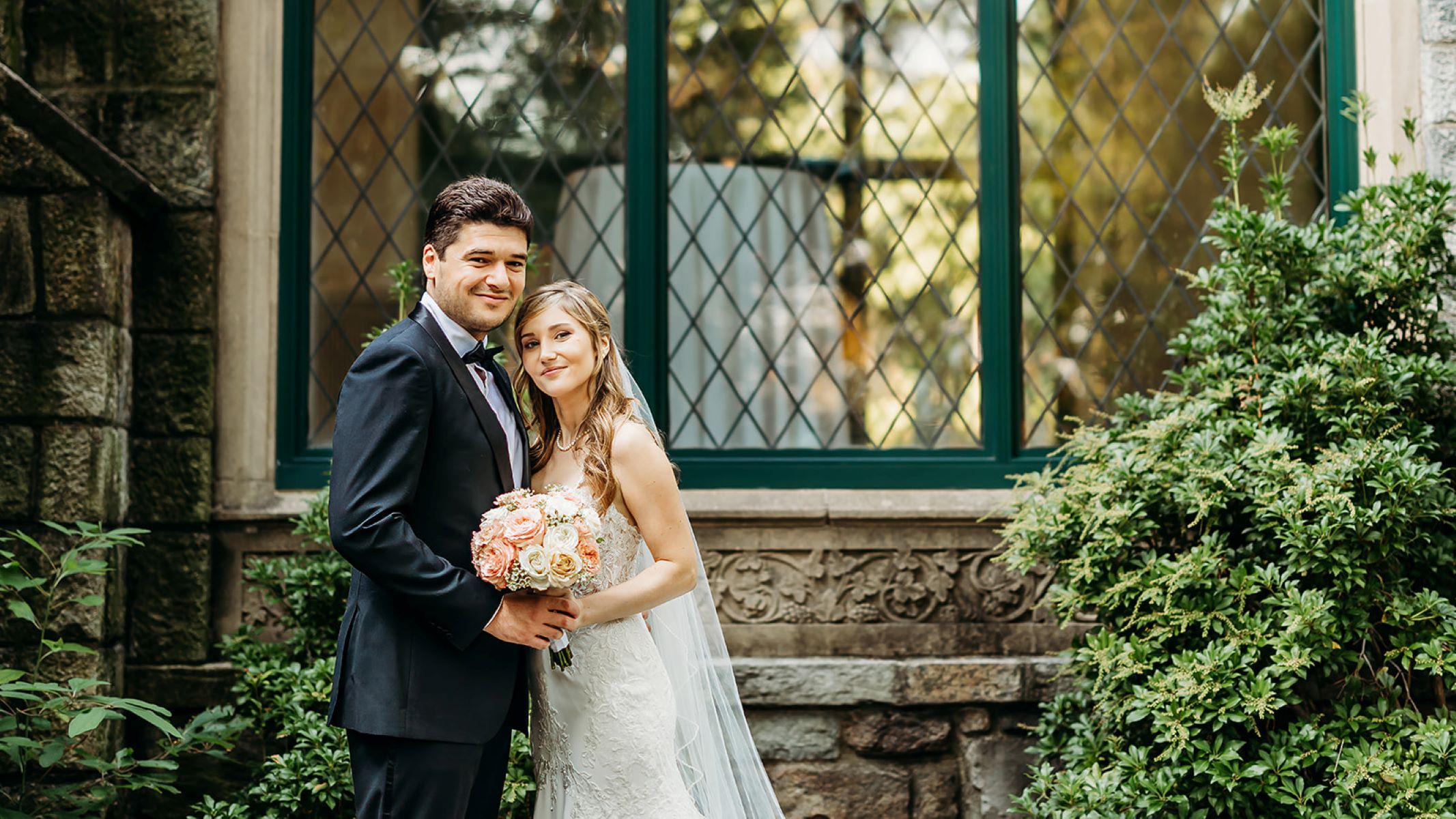 A woman in a white wedding dress with a white wedding veil and a pearl necklace is holding a bouquet of white and pink fowers and smiling and her husband is wearing a black suit and behind them is a gothic style building made of stone and green plants and shrubs 