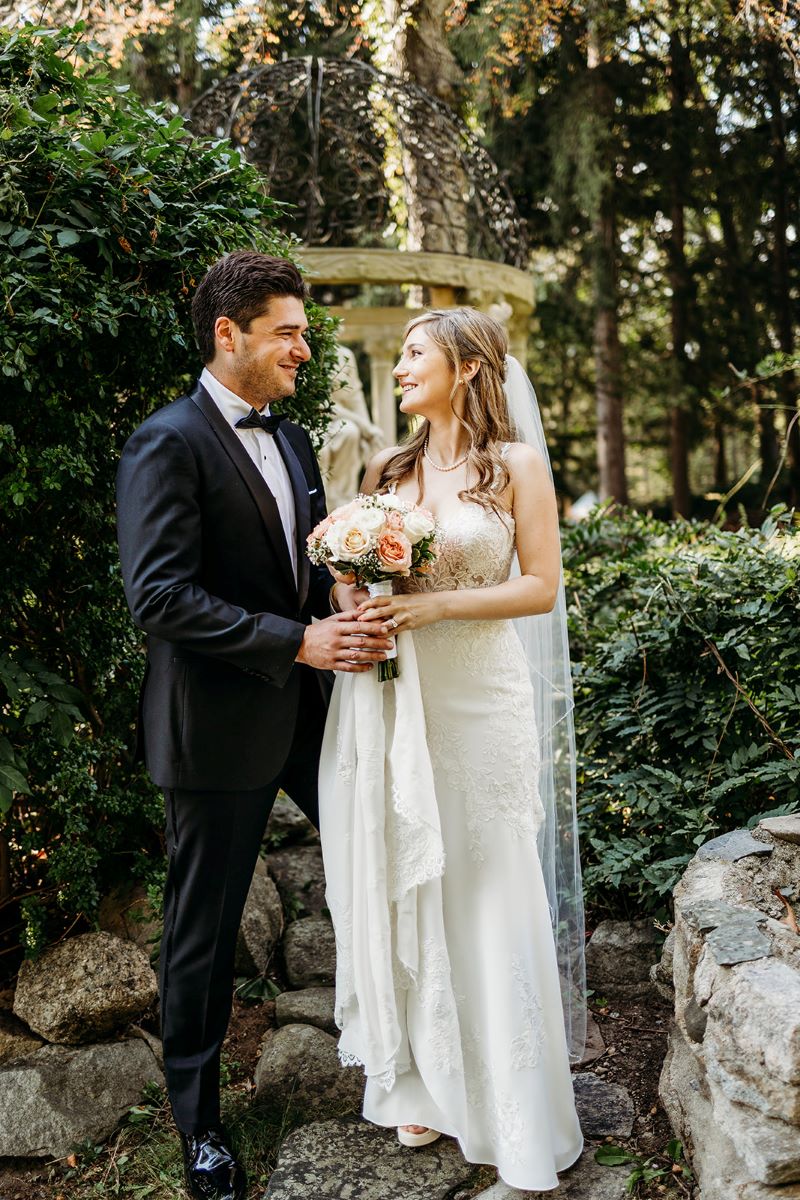 A woman in a white wedding dress with a white wedding veil is holding a bouquet of white and pink flowers and is looking at her husband and smiling and her husband is wearing a black suit and smiling and looking at her behind them is a marble statue and green plants and shrubs and trees