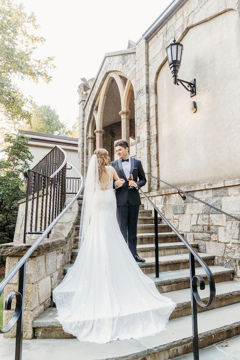 A woman in a white wedding dress and a white veil is standing on the stairs and looking at her husband who is wearing a black suit and is holding a wine glass in his hand behind them is a stone building with gothic architecture 