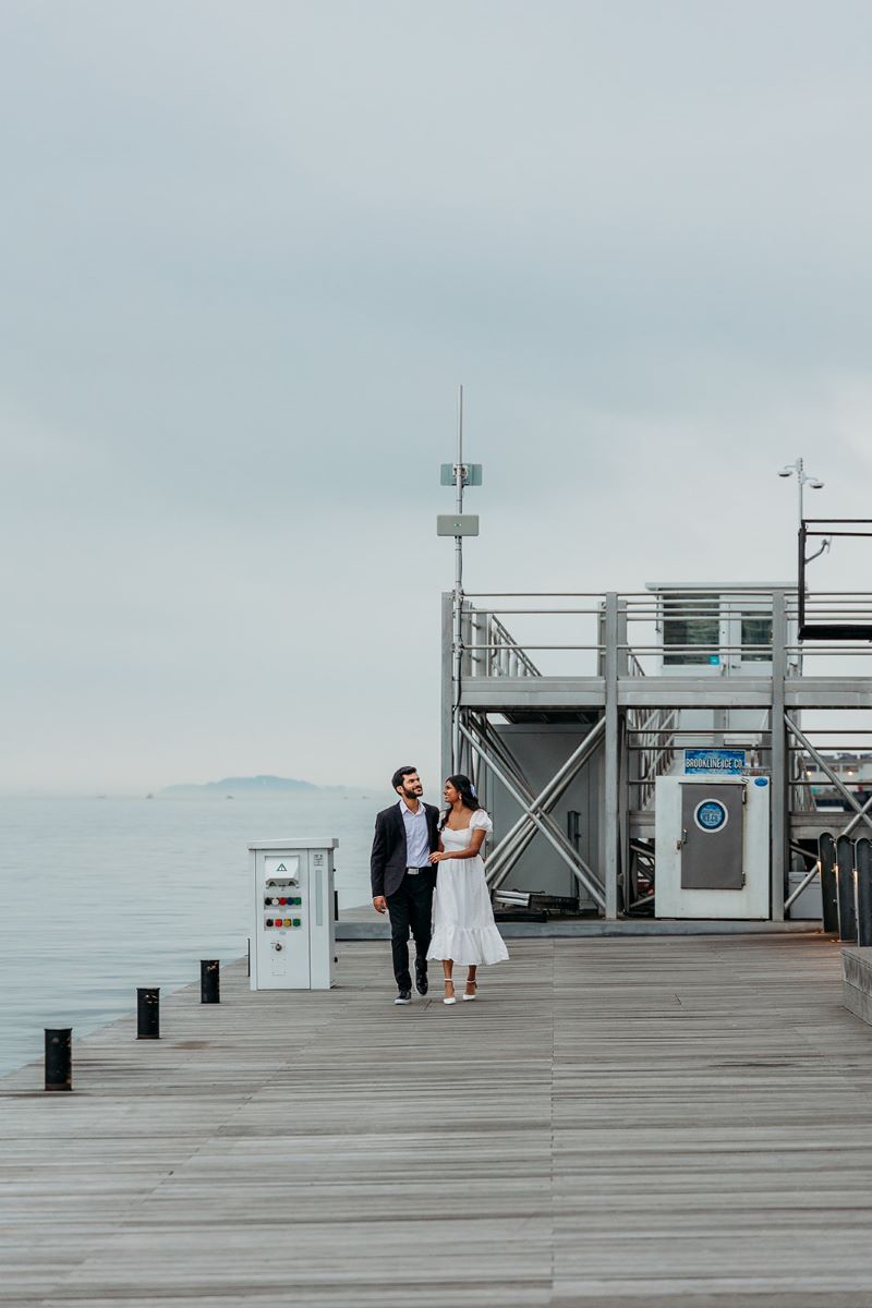 A woman in a white dress and white heels and a man in a suit are walking on a pier and looking at each other they are surrounded by water 