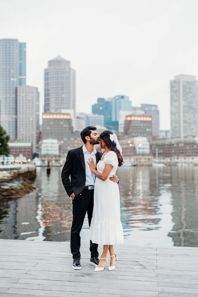 A woman wearing a white wedding dress, a bow, and white heels is touching the chest of her partner who is wearing a black suite and a white button up and is kissing her on the forehead and behind them is water and a city skyline 