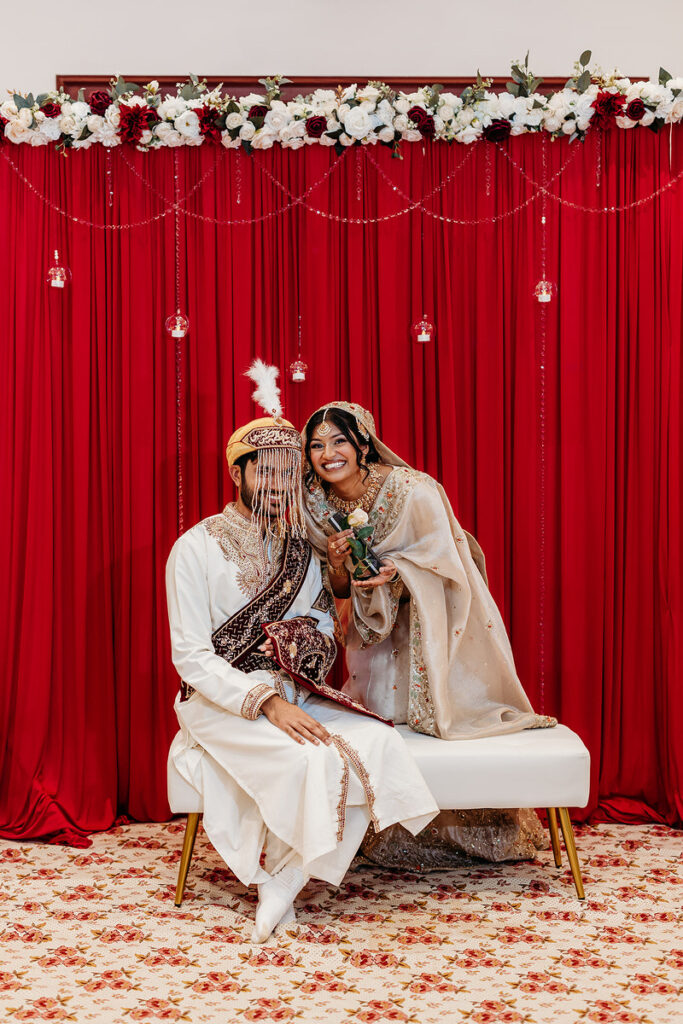 A newlywed couple sitting on a small stool in front of a red drape 