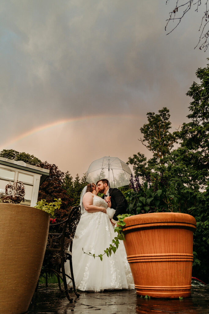 A couple under a clear umbrella kissing with a rainbow behind them 