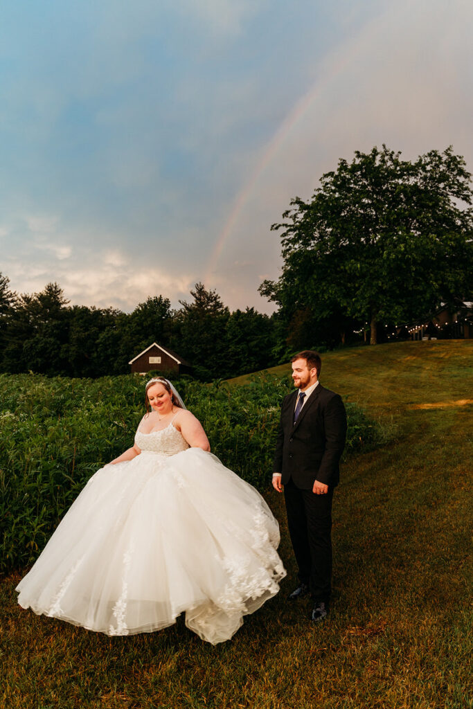 A newlywed couple walking in a field together with a rainbow behind them 
