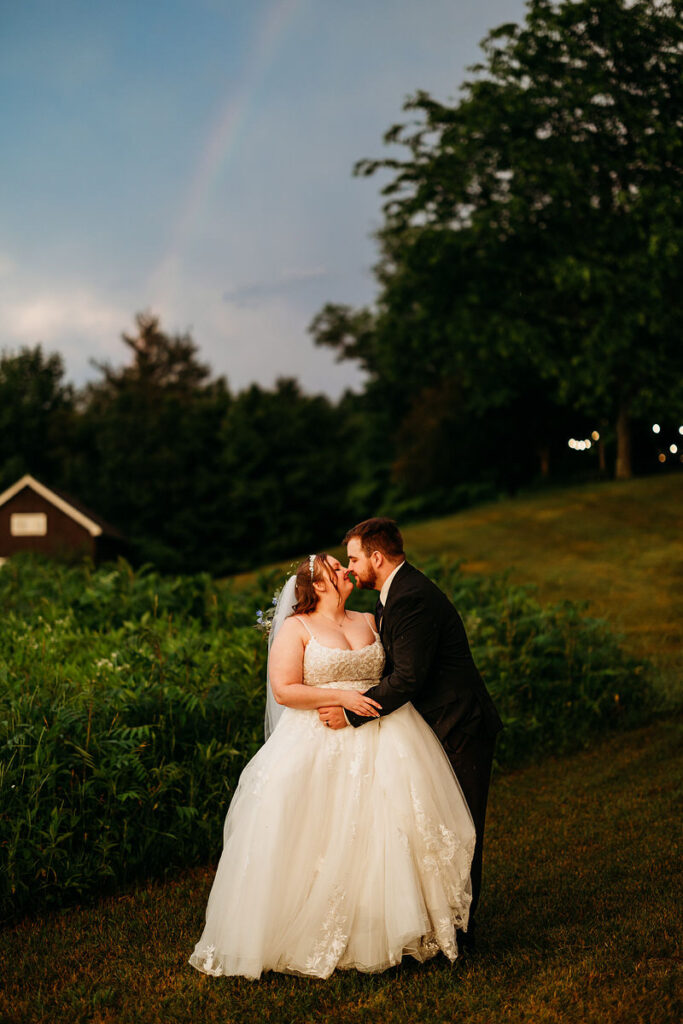 Newlyweds kissing in a field with a rainbow in the sky behind them 
