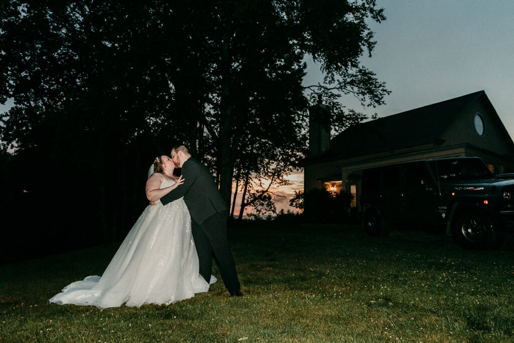 A newlywed couple kissing at dusk in front of a New England style home 