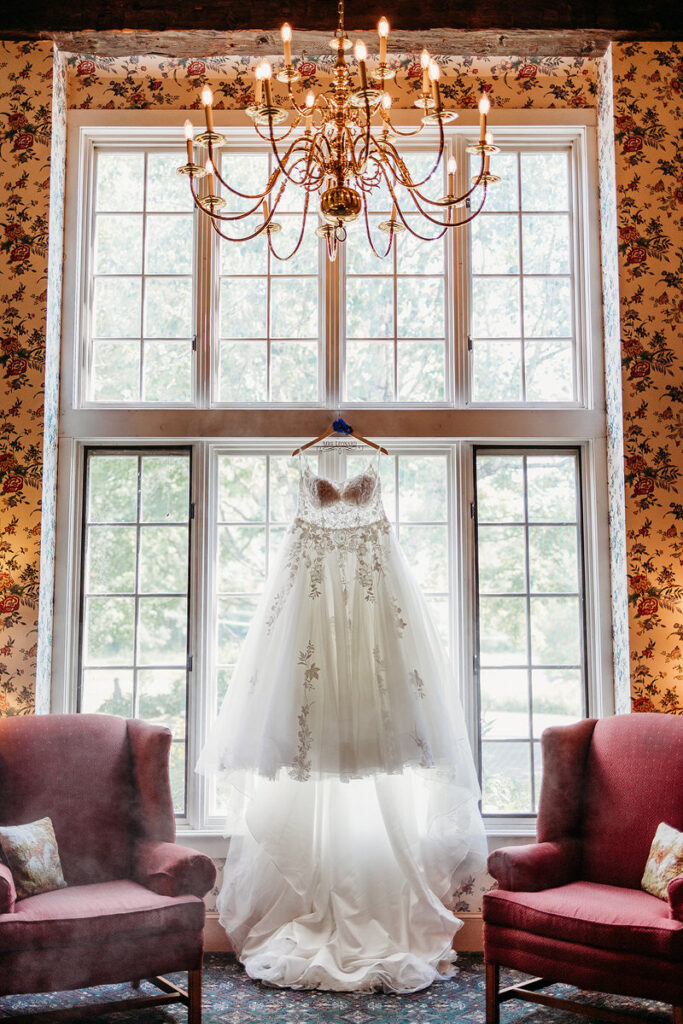 A wedding dress hanging from a grand window with a chandelier in front of it 