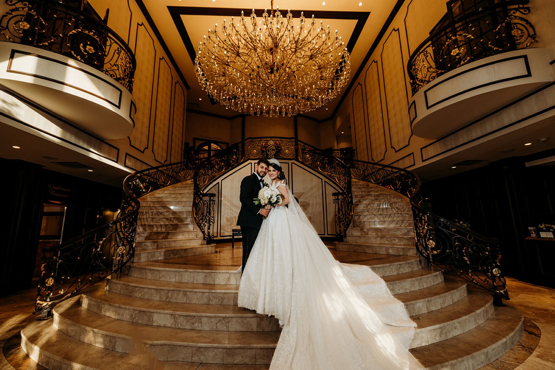 A couple on a grand marble staircase underneath a crystal chandelier the woman is wearing a white wedding dress and veil and tiara and holding a bouquet of flowers and the man is wearing a black tuxedo 