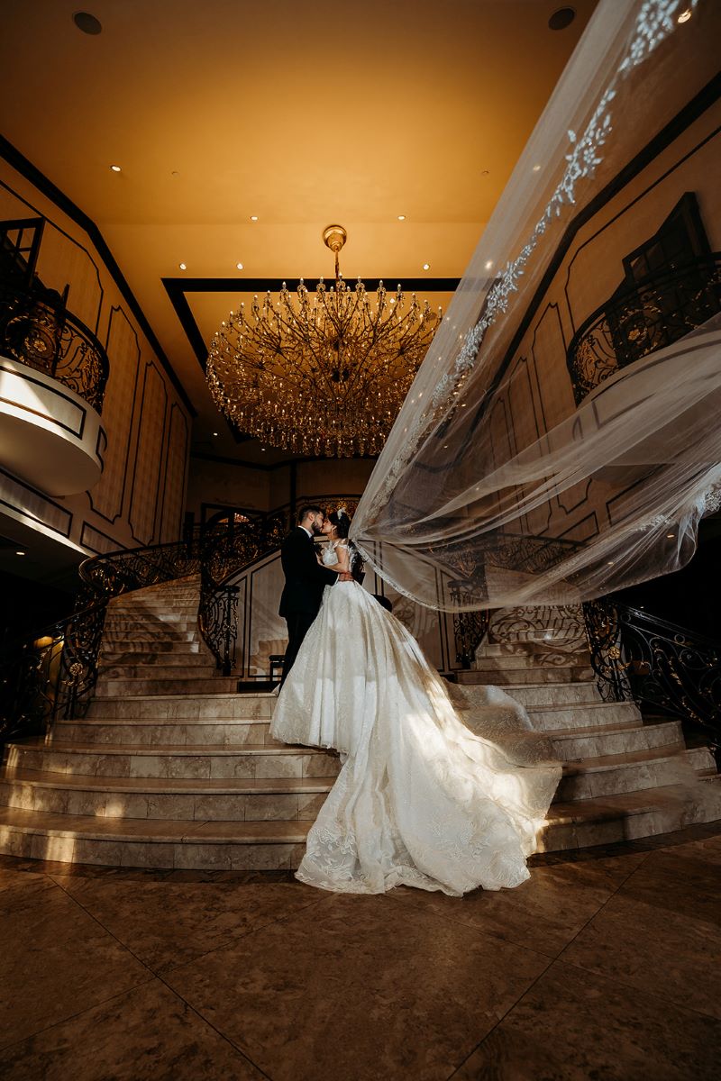 A couple is standing together and kissing on a grand marble staircase underneath a chandelier the woman's veil is floating back behind her and she is wearing a white wedding dress and a tiara and the man is wearing a black tuxedo 