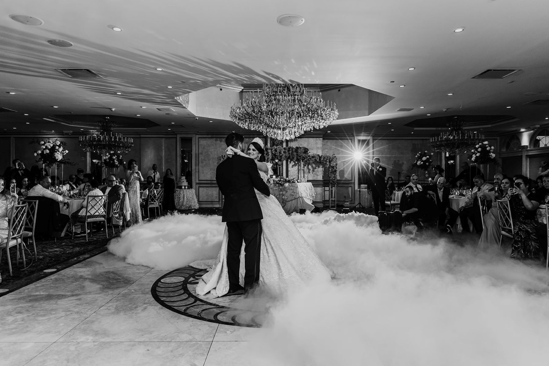 A couple having their first dance at their wedding reception there is a cloud fog machine that makes it looks like they are dancing on top of clouds the guest are sitting and watching the first dance they are dancing underneath a large crystal chandelier