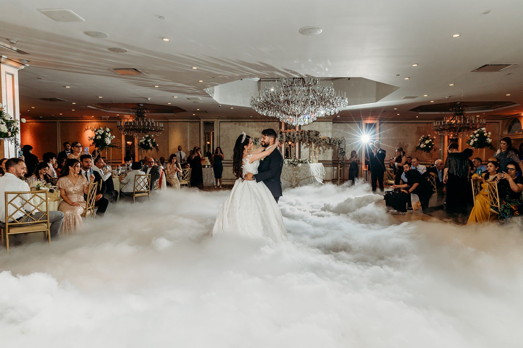 A woman in a white wedding dress wearing a tiara witha man in a black suit they are both dancing and there is a cloud fog machine that makes it appear they are dancing on top of white puffy clouds and hanging overhead is a large crystal chandelier while their wedding guests watch their first dance 