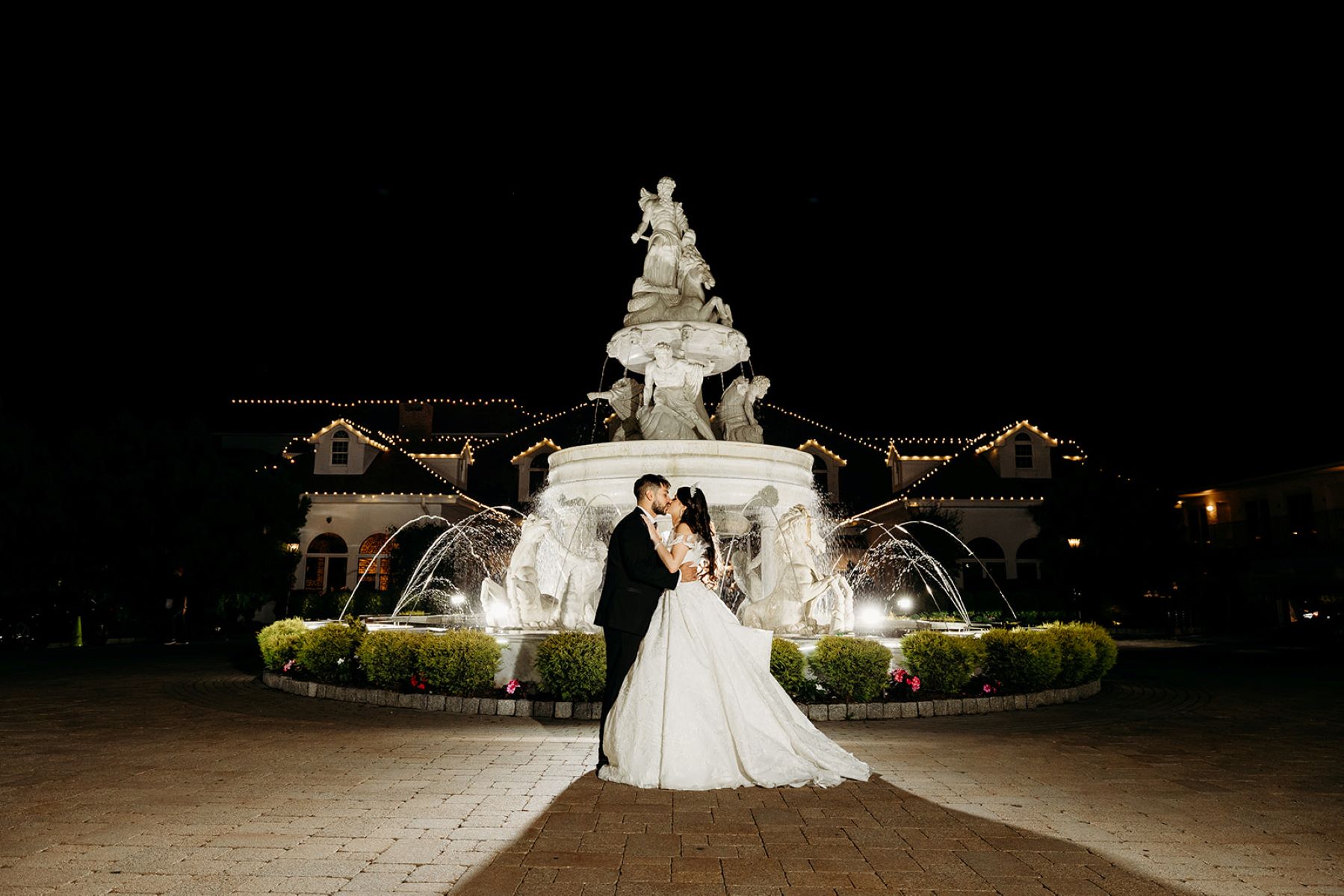 A woman wearing a white wedding dress kissing her partner who is wearing a black tuxedo they are kissing at night in front of a lit up fountain that also has various statues 