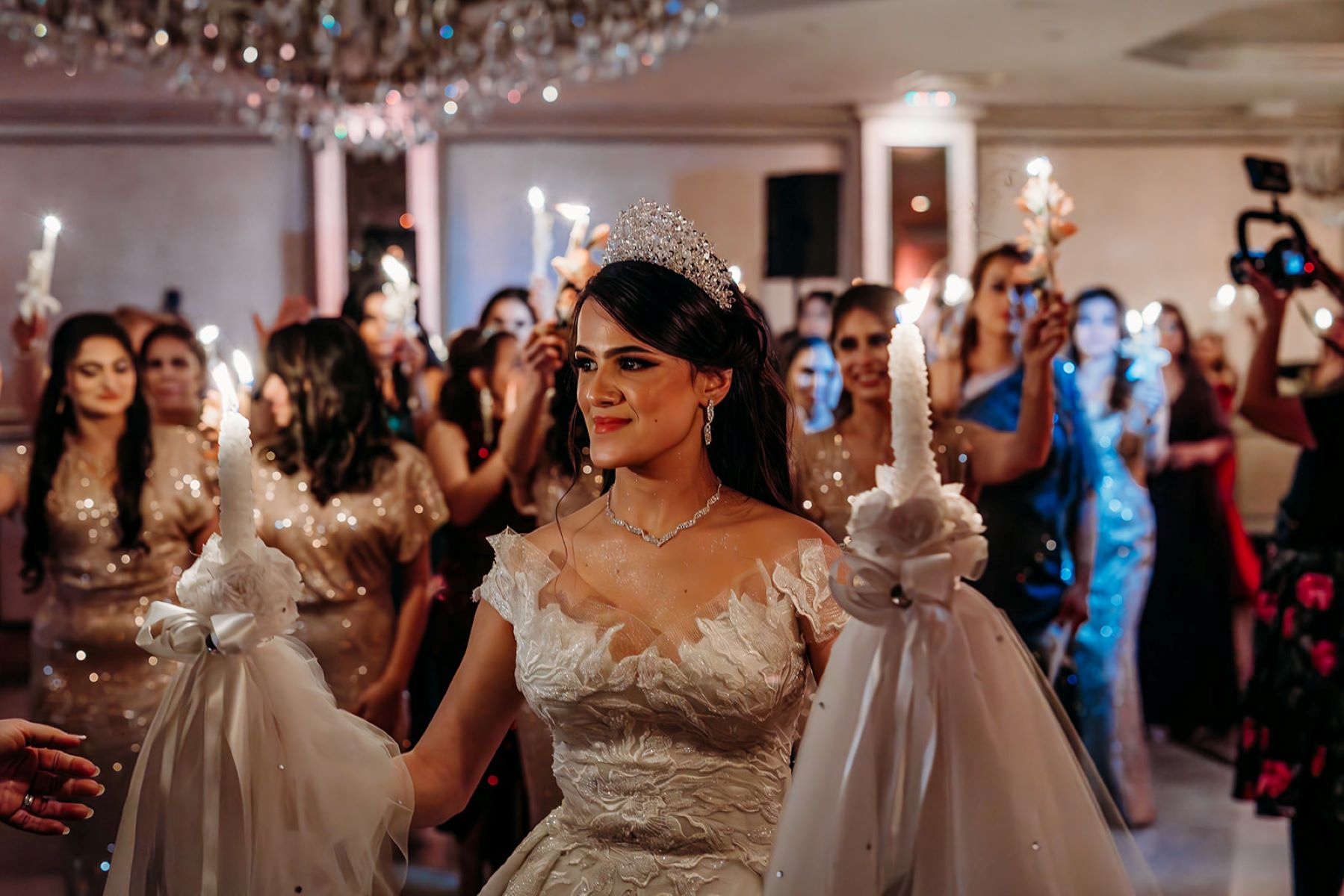 A woman in a white wedding dress holding two candles in her hands and who is wearing a tiara and behind her are other women holding candles 