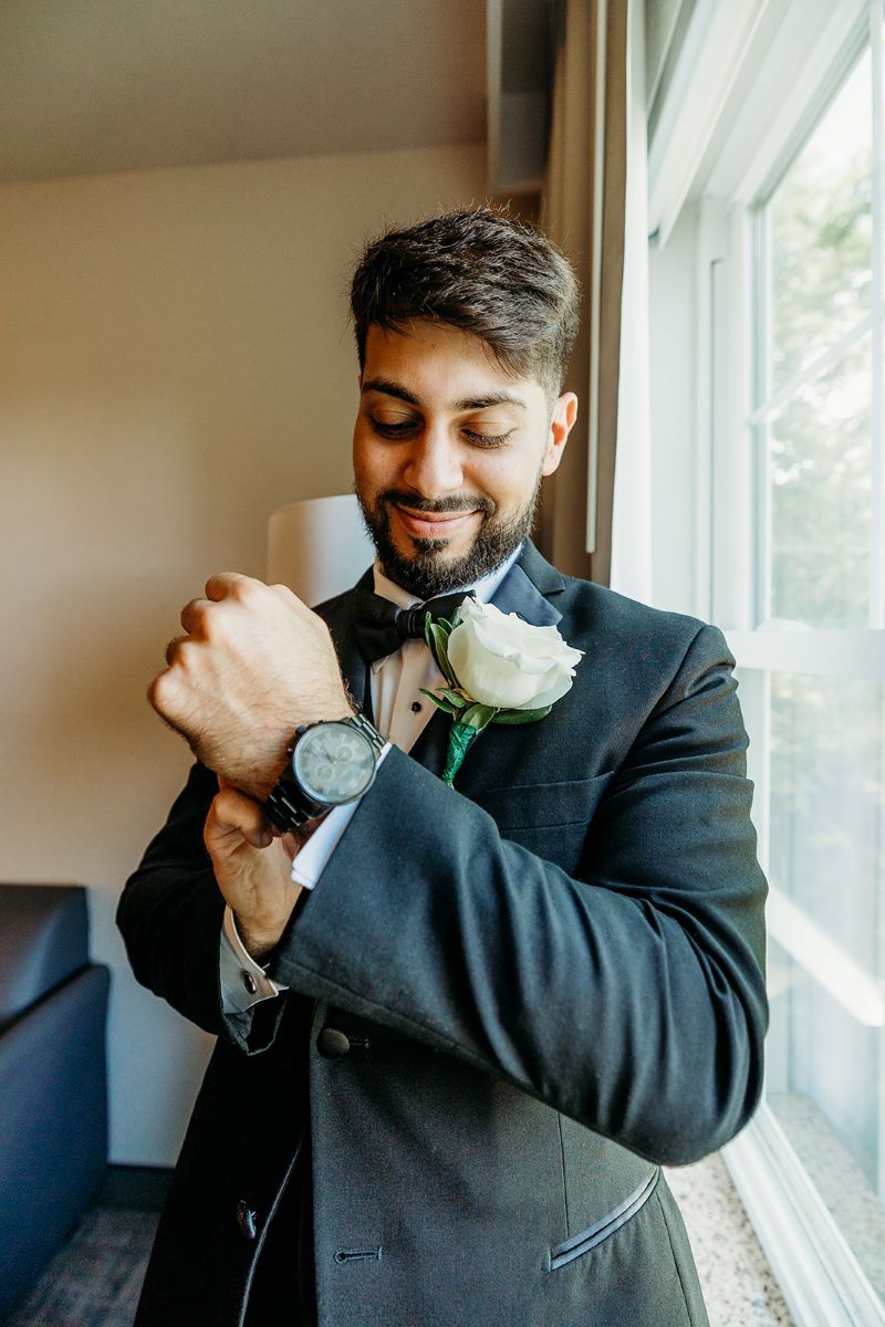 A man is getting ready for his wedding he is putting on his watch and wearing a black suit and black bowtie 