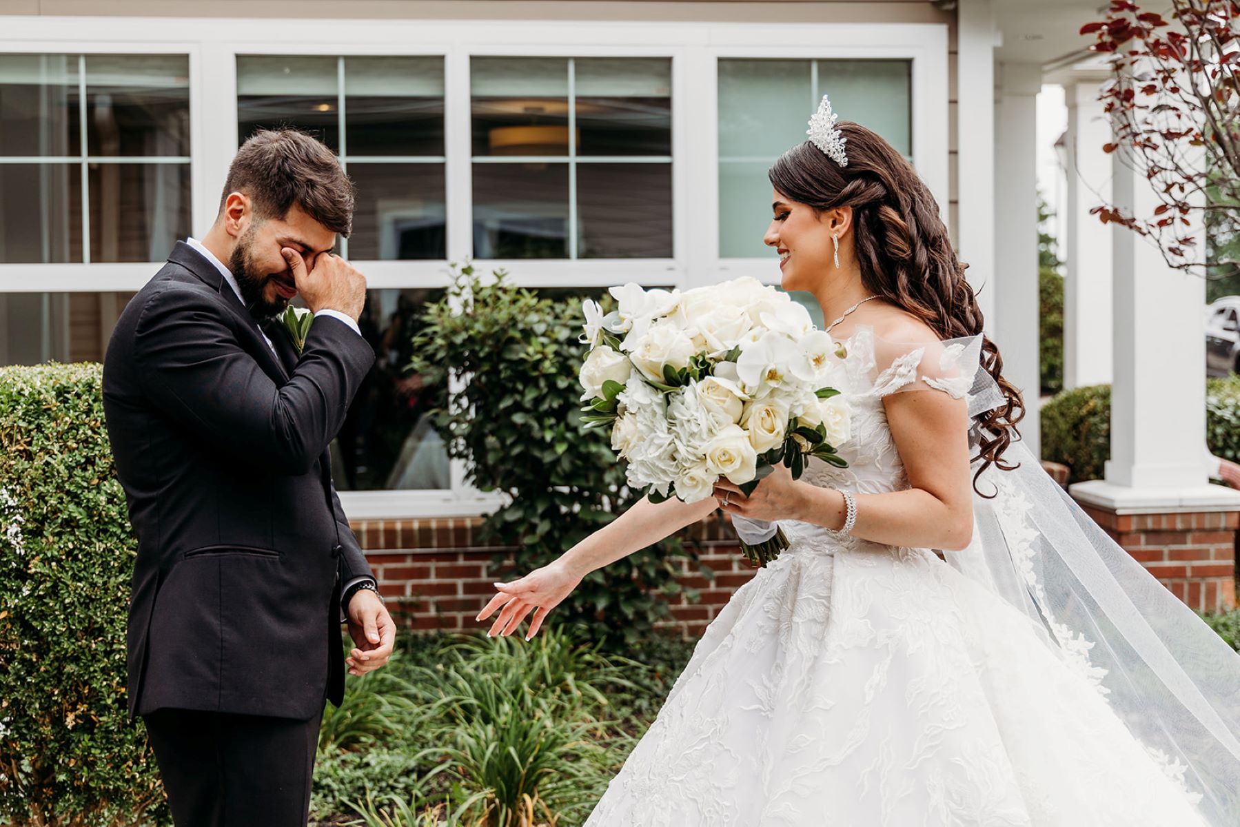 A man wearing a black tuxedo tearing up when he sees his partner in her white wedding dress his partner is also wearing a tiara and holding a bouquet of white flowers