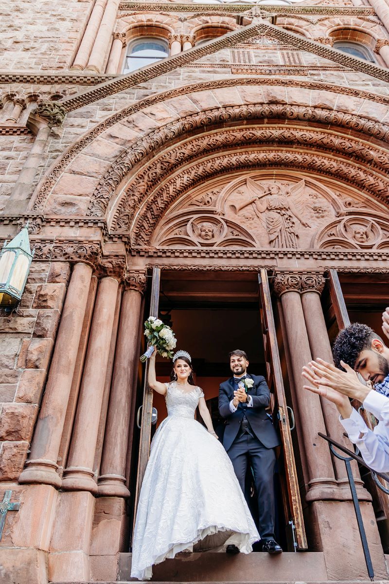 A couple leaving their wedding ceremony from a church the woman is wearing a white wedding dress and a tiara and is holding up her bouquet of flowers and the man is wearing a black suit and clapping his hands together 