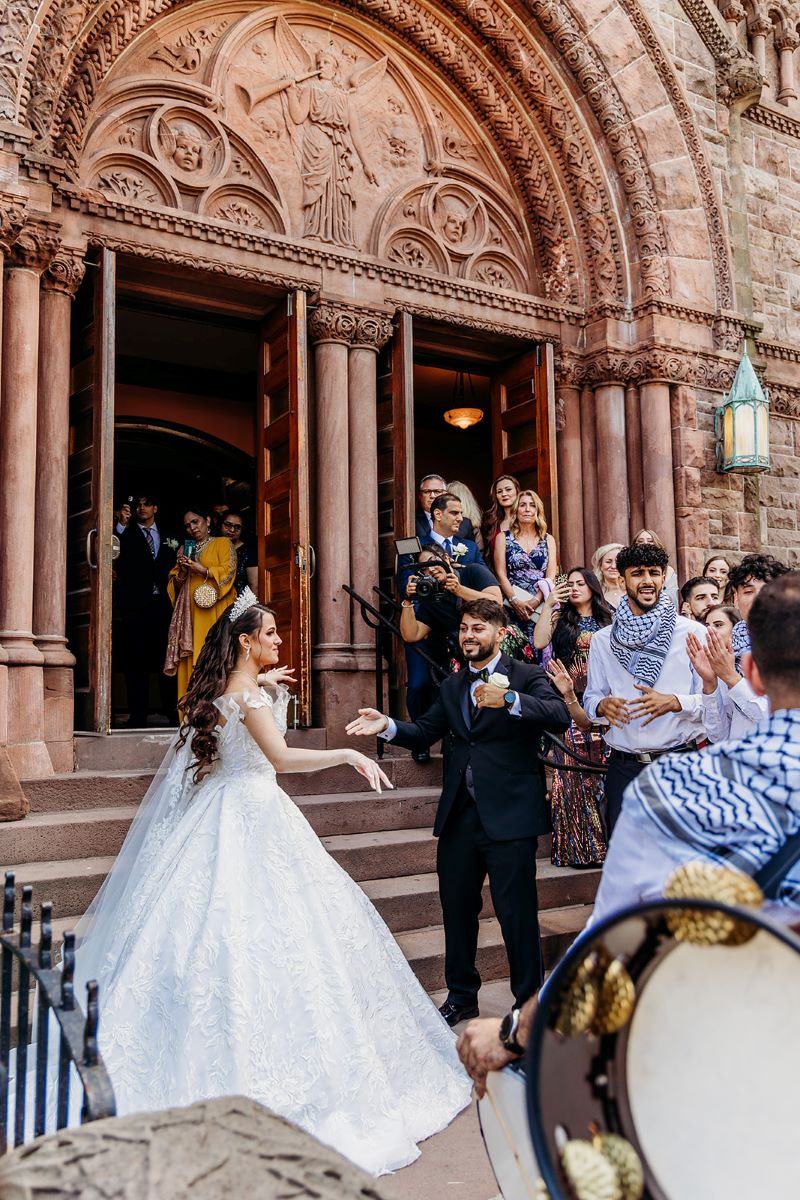 A couple dancing in front of church steps after their wedding ceremony while their guests cheer for them the woman is wearing a white wedding dress and a tiara and the man is wearing a black suit 