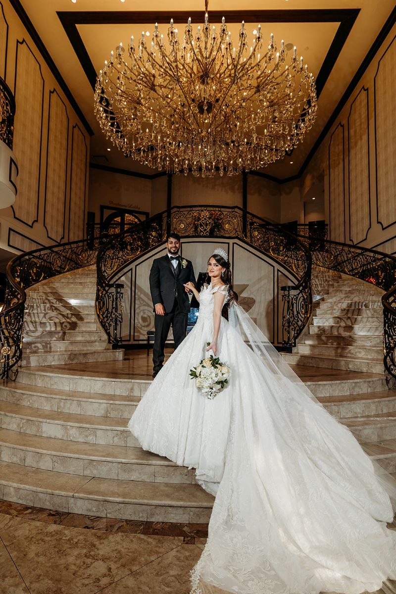 A couple is standing on a grand marble staircase underneath a large crystal chandelier and the woman is wearing a white wedding dress and has on a white veil and she is holding the hand of her partner with her other hand she is holding a bouquet of flowers the man is wearing a black tuxedo and is standing behing her 