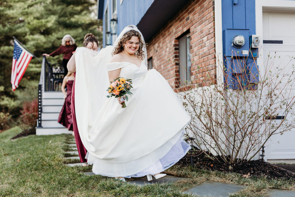 A person in a wedding dress walking along stones in grass as someone holds up the back of their dress 