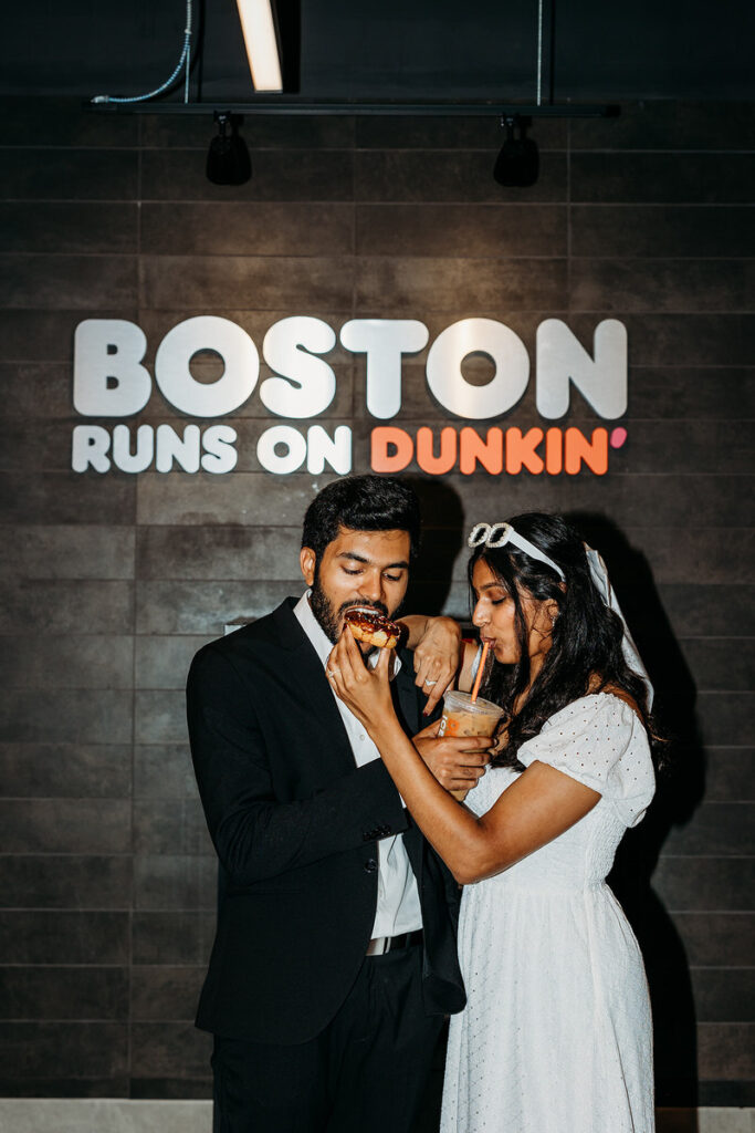 A newlywed couple standing in front of a Dunkin sign while one feeds the other a donut and one holds a drinkn to the other's lips 