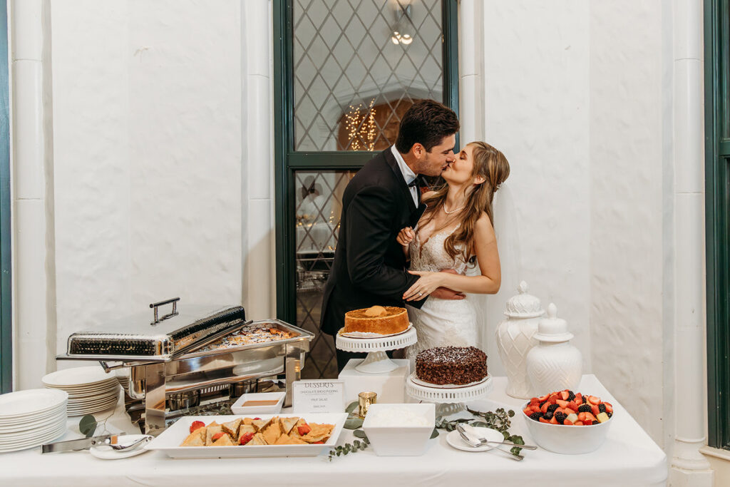 A newlywed couple kissing and standing in front of a table of desserts at their reception